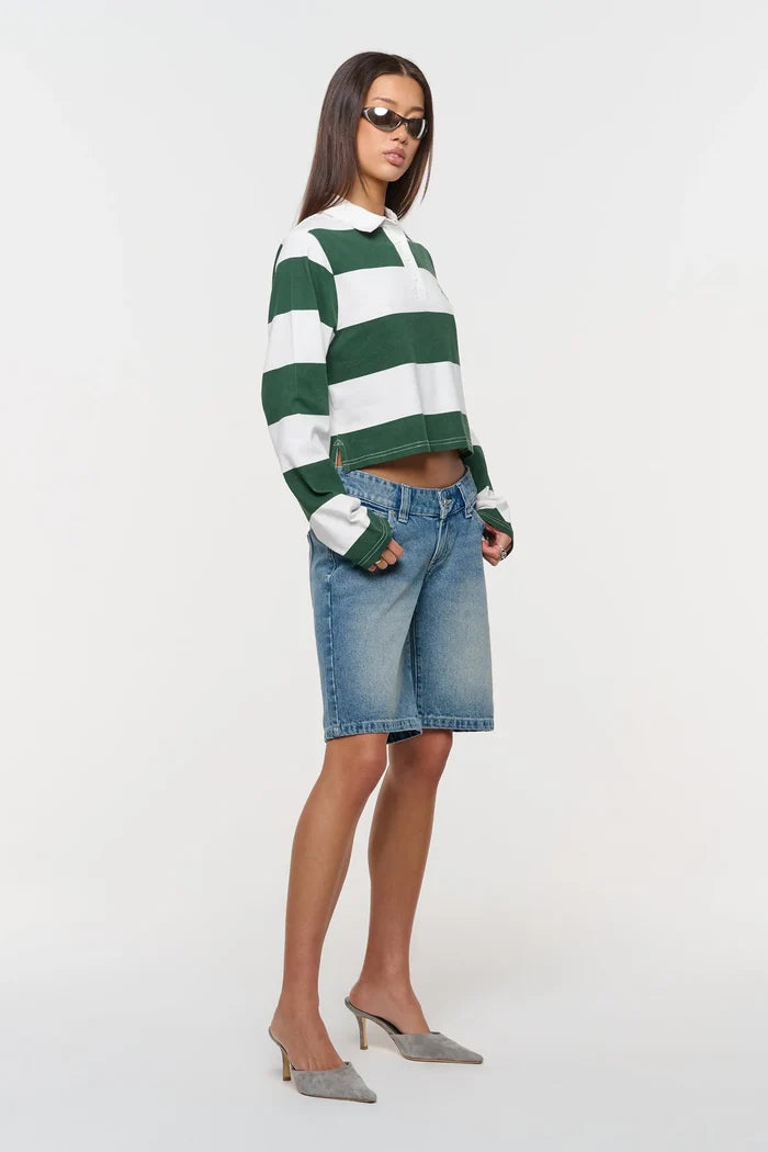 A model wearing a green and white striped long-sleeve crop top, blue denim shorts, and gray pointed-toe mule heels, standing against a plain white studio background.
