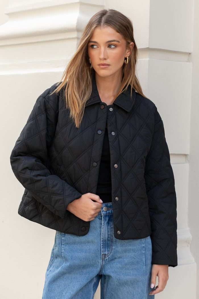 Woman wearing a black jacket and blue jeans standing against a white building.