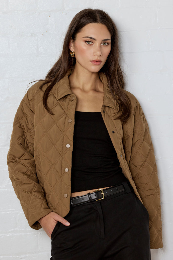 Woman wearing a brown quilted jacket over a black top and pants against a white brick wall.