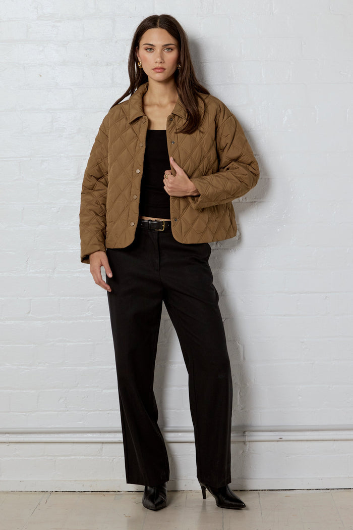 Woman wearing a brown jacket and black pants against a white brick wall.