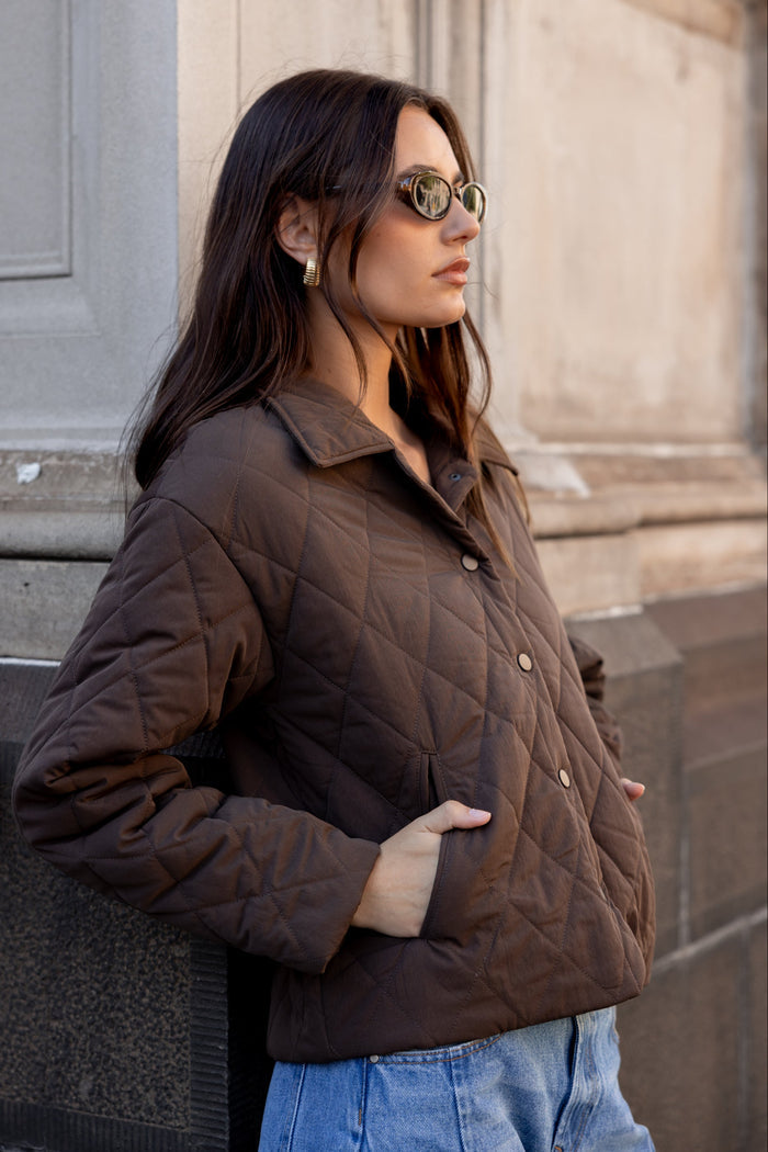 Woman wearing a brown quilted jacket and sunglasses against a building background