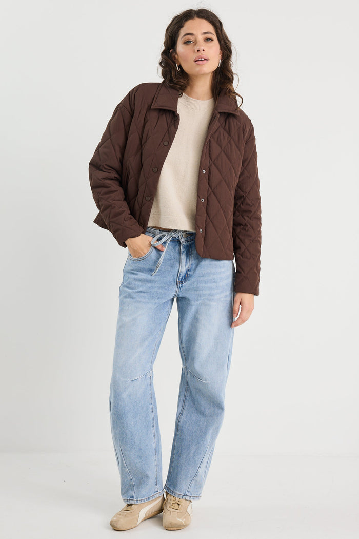 Woman wearing a brown jacket, beige shirt, and blue jeans on a white background