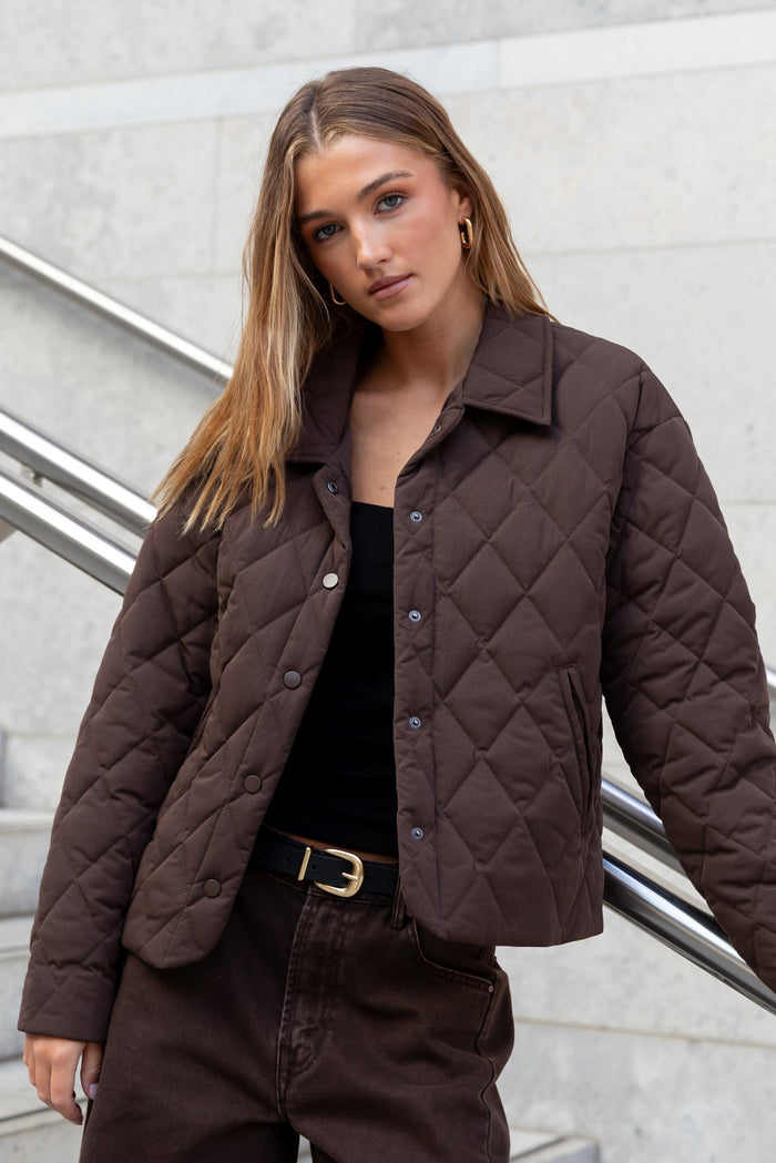 Woman wearing a brown quilted jacket standing on a staircase with a light gray wall background