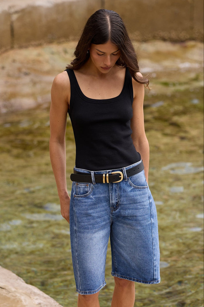 A woman standing on a stone ledge next to a shallow stream, wearing a black tank top, blue denim cutoff shorts, a black belt with a gold buckle, and black flip-flops.