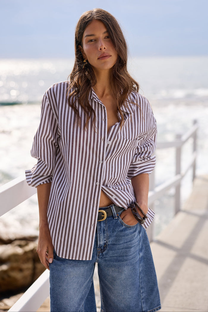 A woman wearing a brown and white vertically striped button-down shirt and blue denim jeans, standing on a sunny boardwalk with the ocean in the background.