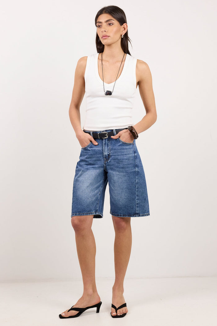 A model stands against a plain white studio background wearing blue denim shorts, a white sleeveless top, black sandals, and layered necklaces.