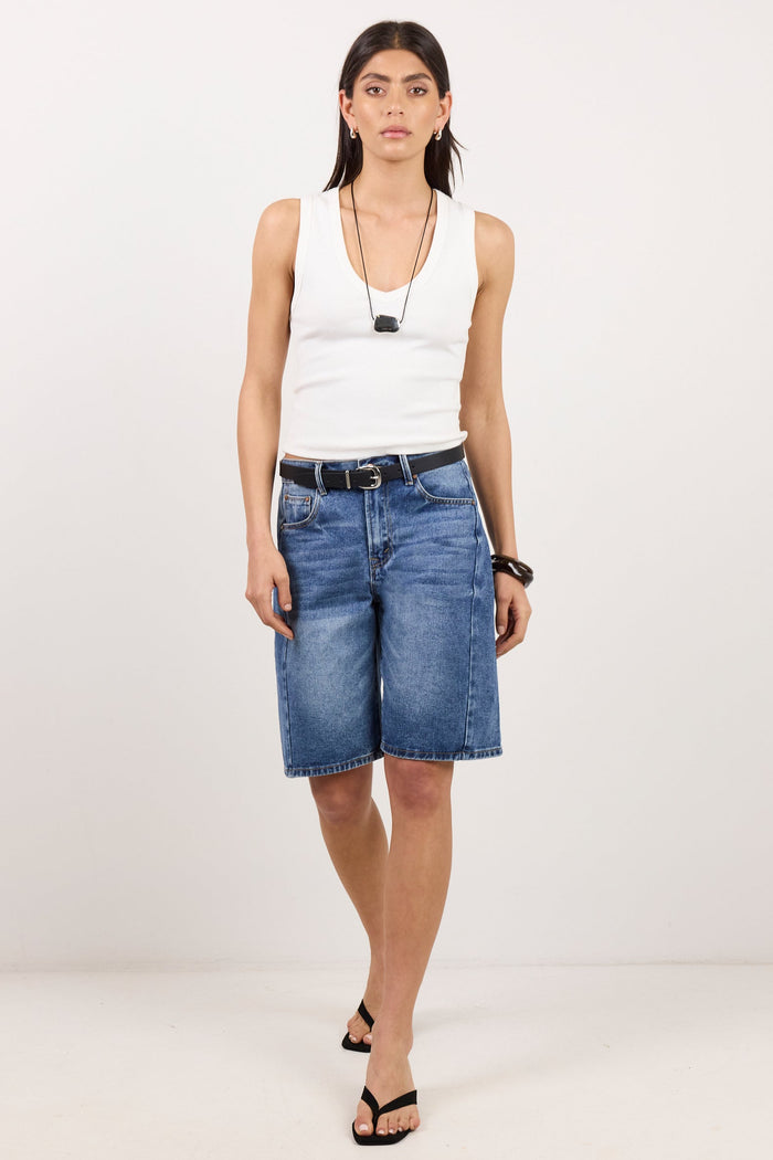 A woman modeling a pair of medium-wash blue denim shorts with a black belt, paired with a white sleeveless top and black sandals, standing against a plain white studio background.
