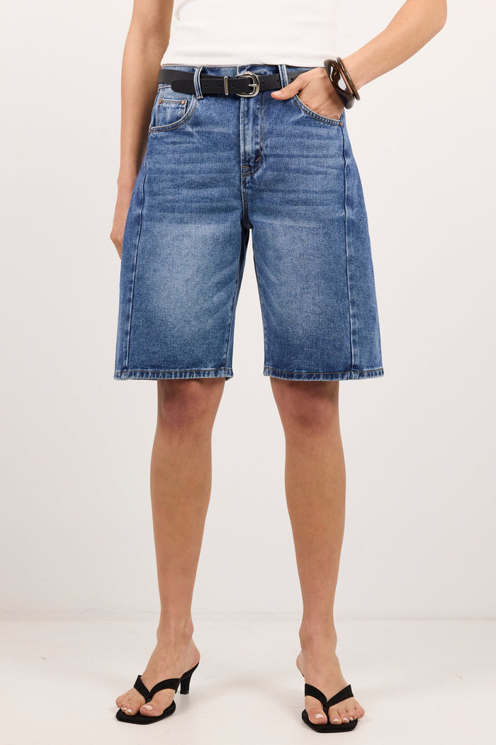 A model wearing medium-wash blue denim shorts with a black belt, paired with a white top and black heeled sandals, standing against a plain white studio background.