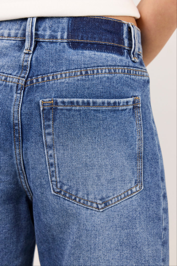 Close-up of the back waistband and right back pocket of blue denim jeans, worn by a model in a studio setting.
