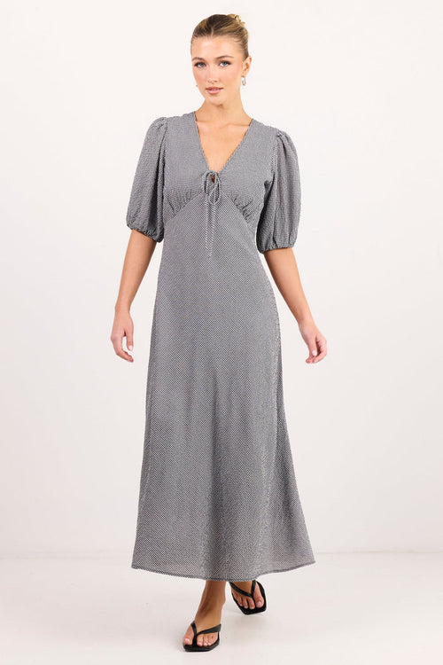 A woman modeling a long gray midi dress with puff sleeves, a V-neckline, and a tie detail at the bust, photographed against a plain light studio background.