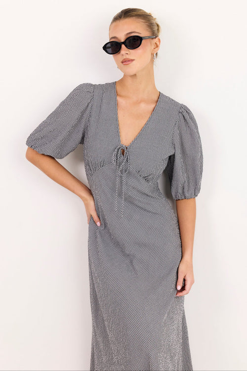 A woman modeling a gray and white micro-patterned midi dress with puff sleeves, a V-neckline, and a front tie detail, set against a plain white background.