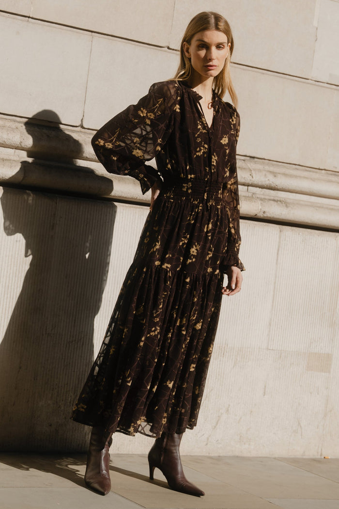 Woman in a dark patterned dress standing against a light stone wall.