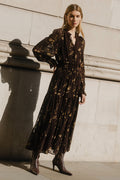 Woman in a dark patterned dress standing against a light stone wall.