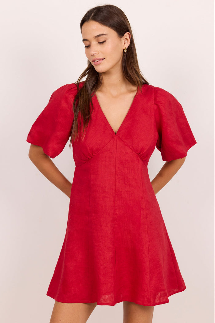 A woman modeling a red short puff-sleeve dress with a V-neckline, photographed against a plain light beige studio background.