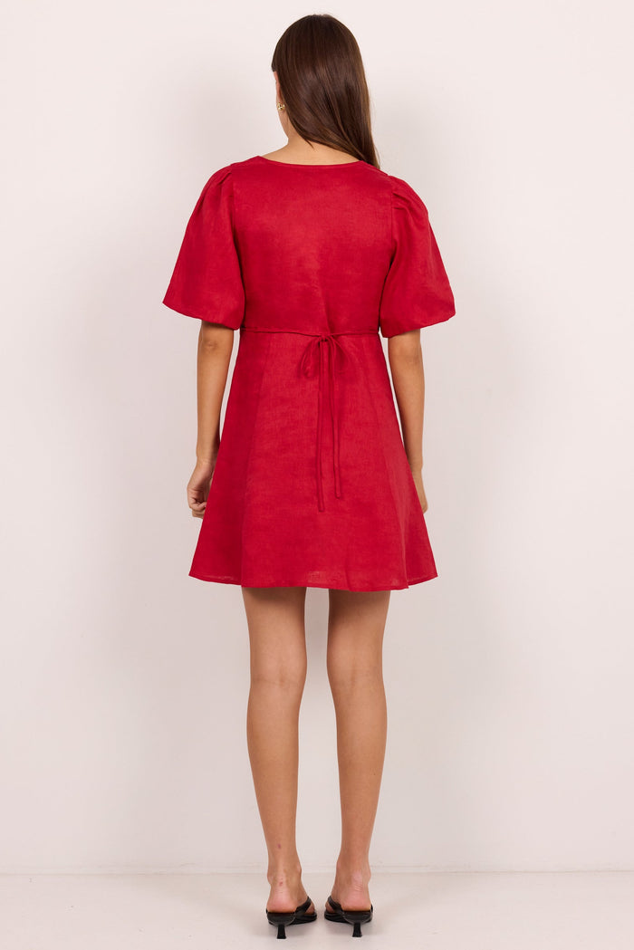 A woman viewed from the back wearing a short-sleeve red dress with a tie waist, standing against a plain light background.