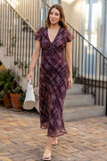 A woman wearing a dark purple and brown plaid midi dress with flutter sleeves walks down a brick courtyard path next to a building with black metal stairs.