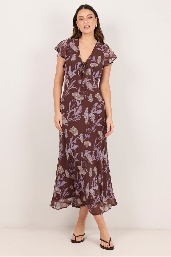 A woman modeling a brown midi dress with a purple floral pattern, ruffled cap sleeves, and a V-neckline, standing against a plain off-white studio background.