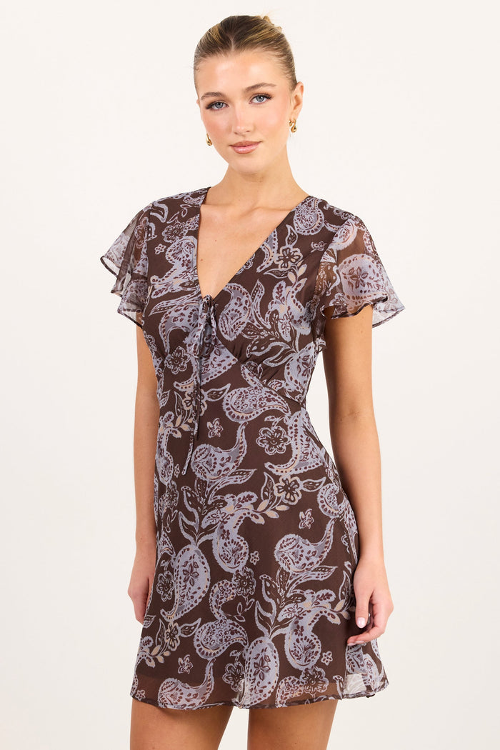 A woman modeling a short brown mini dress with a white paisley pattern, flutter sleeves, and a V-neckline, photographed against a plain off-white studio background.
