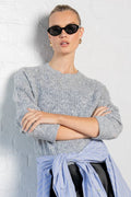 Person wearing a gray sweater, light blue shirt, black skirt, and black shoes against a white wall.