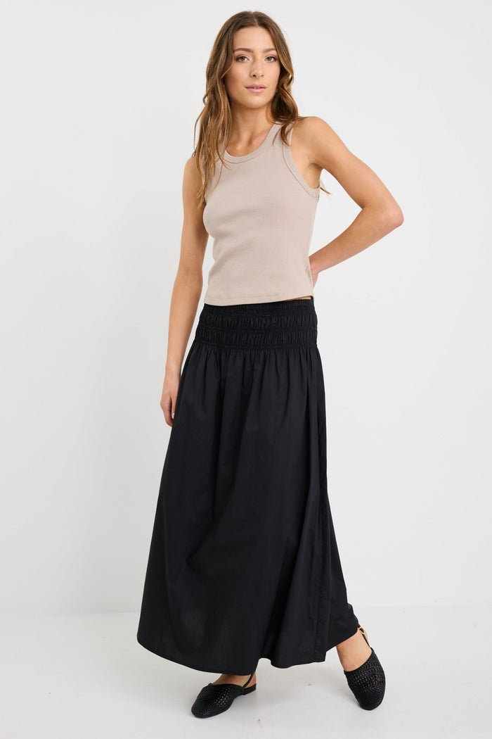Woman wearing a black cotton maxi skirt with shirred waistband, styled with a taupe ribbed tank and black woven flats.