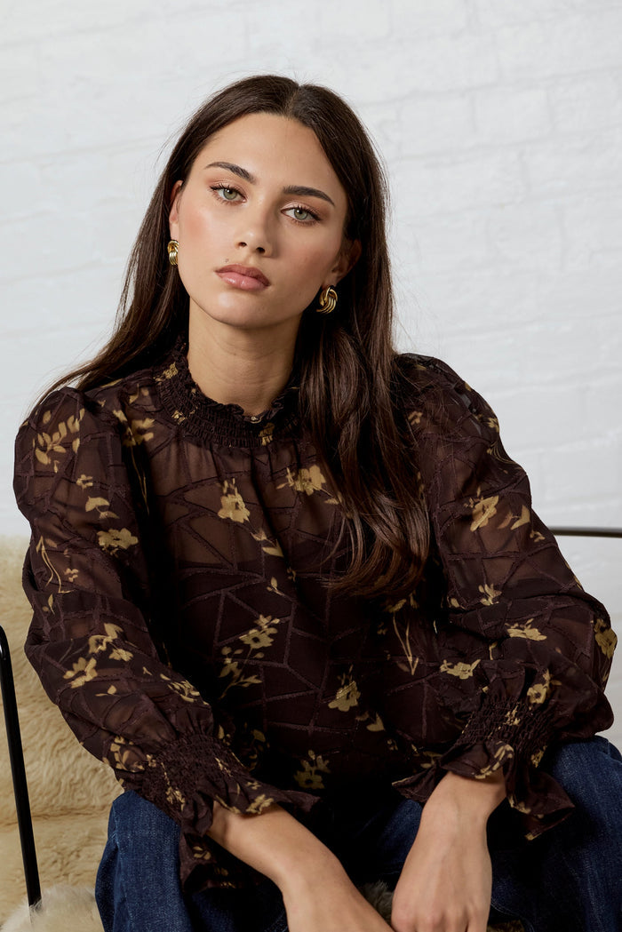 Woman sitting on a chair wearing a dark floral blouse and jeans against a light background