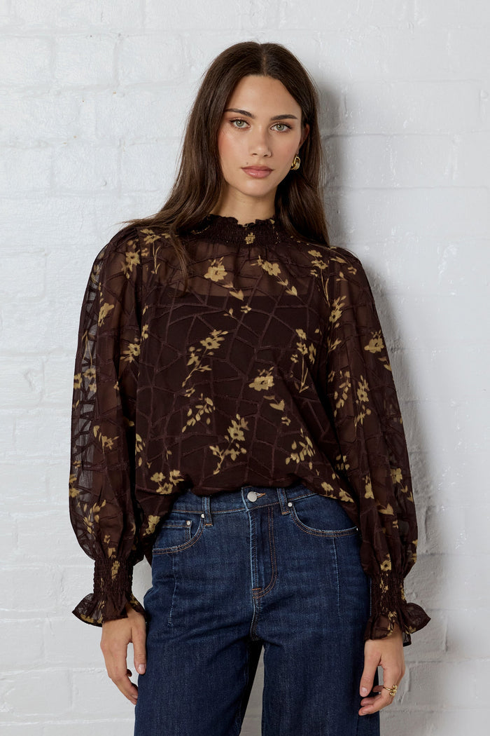 Woman wearing a dark floral blouse and jeans against a white brick wall.