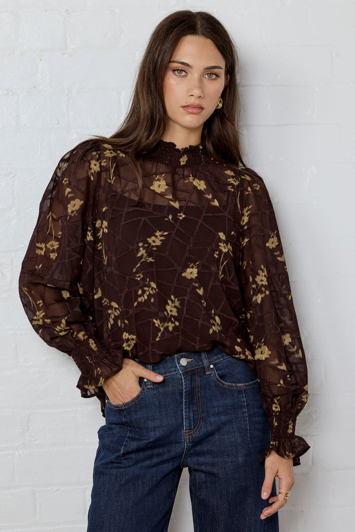 Woman wearing a dark floral blouse and blue jeans against a white brick wall.