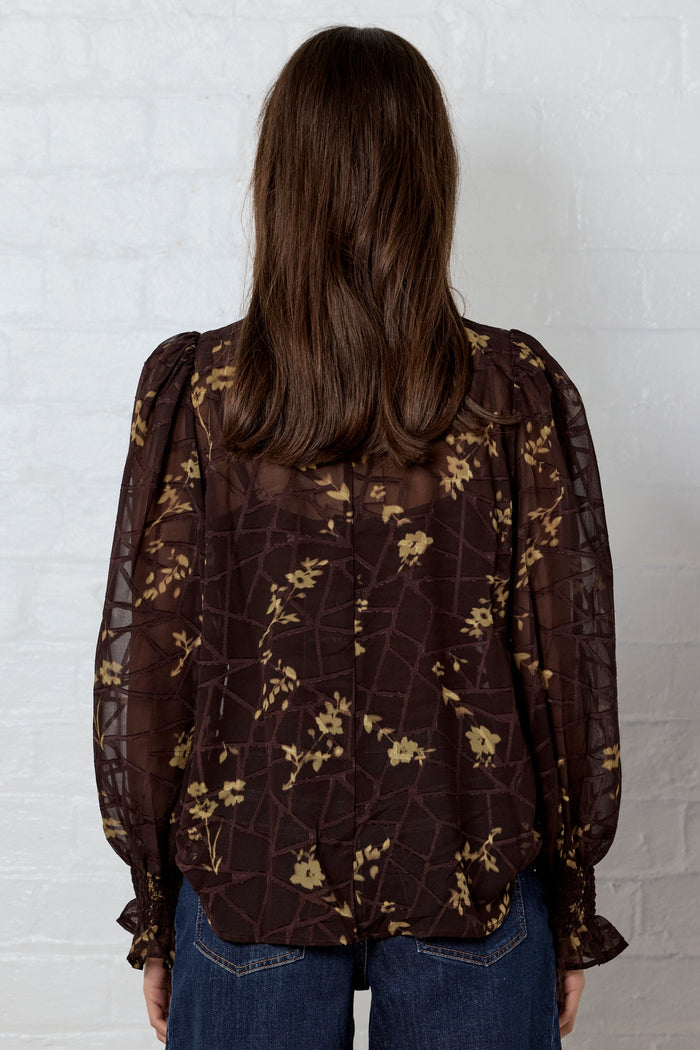 Person wearing a brown floral blouse and blue jeans against a white brick wall.