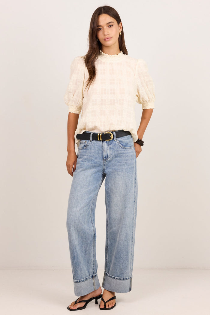 A woman modeling a cream-colored textured puff-sleeve top and light-wash wide-leg blue jeans with a black belt, standing against a plain off-white studio background.
