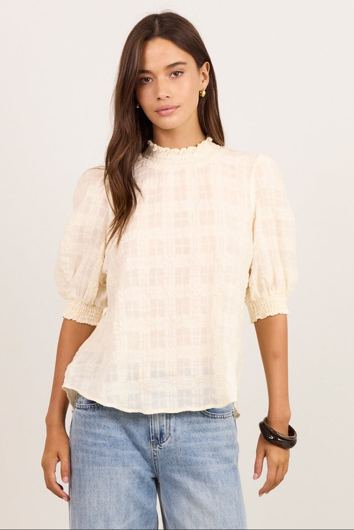 A woman modeling a cream-colored textured puff-sleeve blouse with a subtle grid pattern, paired with light-wash wide-leg jeans and black sandals, standing against a plain off-white studio background.