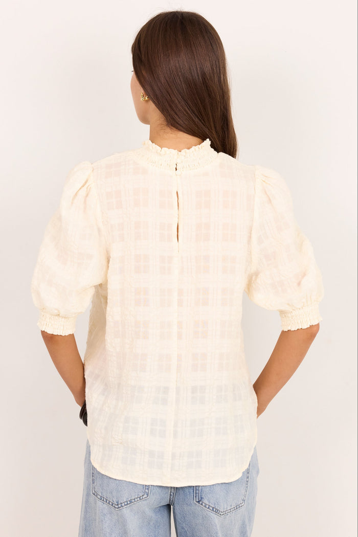 A woman viewed from the back wearing a cream-colored textured blouse with puff sleeves and a high ruffled collar, paired with light blue jeans, against a plain white background.
col_pattern