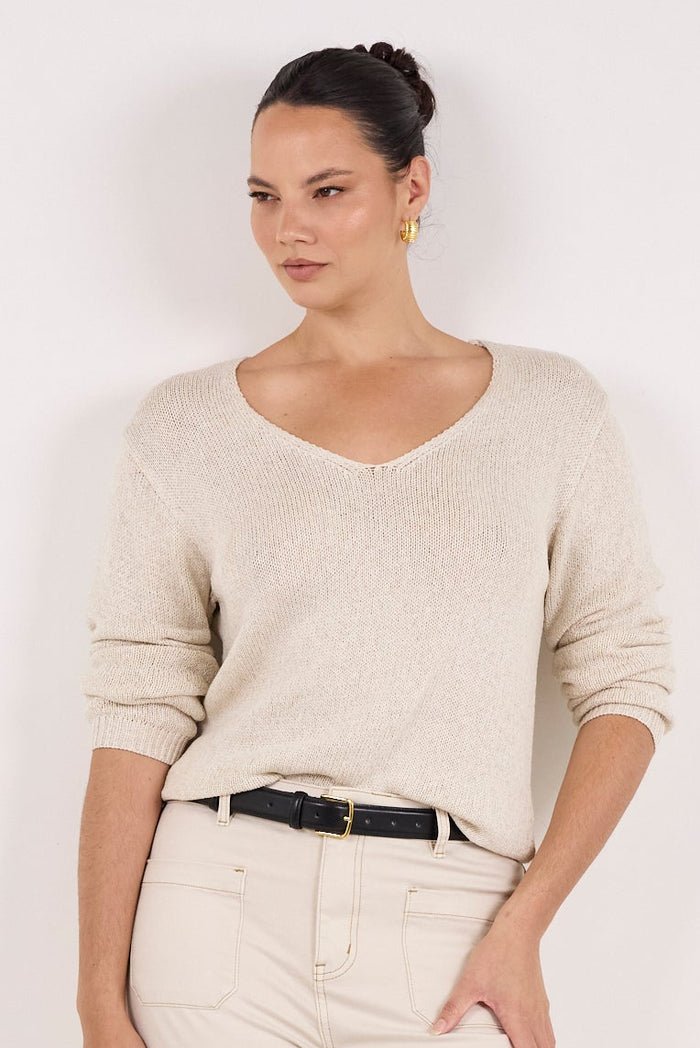 A woman modeling a light beige knit V-neck sweater and matching wide-leg trousers with a thin black belt, paired with black flats, against a plain white studio background.