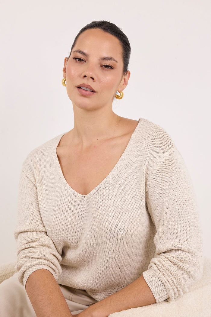 A woman wearing a beige knit V-neck sweater and matching trousers, seated against a plain light background.