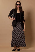 A woman models a black blazer, black top, and a black and white plaid maxi skirt against a solid beige studio backdrop.
