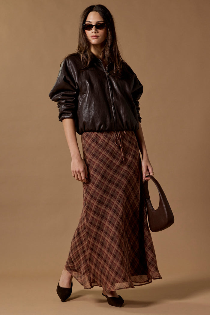 A model wearing a black leather bomber jacket, a brown and red plaid midi skirt, black pointed-toe heels, and carrying a dark brown handbag against a solid beige studio backdrop.