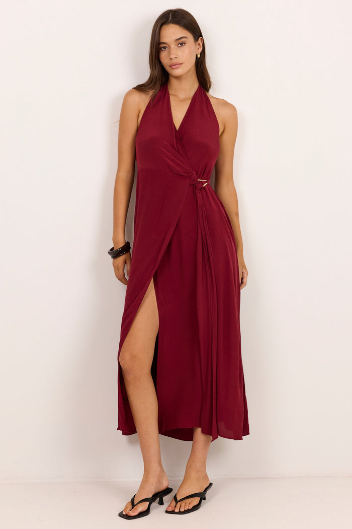 A woman modeling a sleeveless halter-neck midi dress in deep burgundy with a wrap-style front and high side slit, standing against a plain off-white studio background.