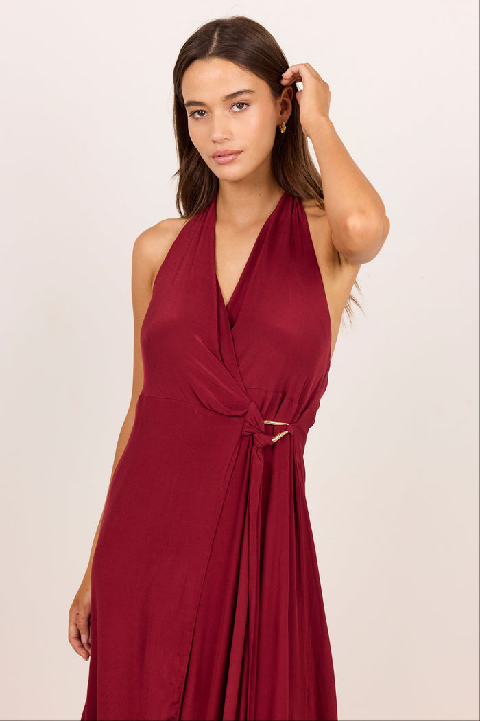 A woman modeling a sleeveless halter-neck maxi dress in deep burgundy with a wrap-style V-neck and a tie detail at the waist, set against a plain light beige studio background.