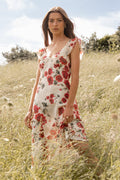 Woman in a floral dress standing in a grassy field with trees and blue sky in the background