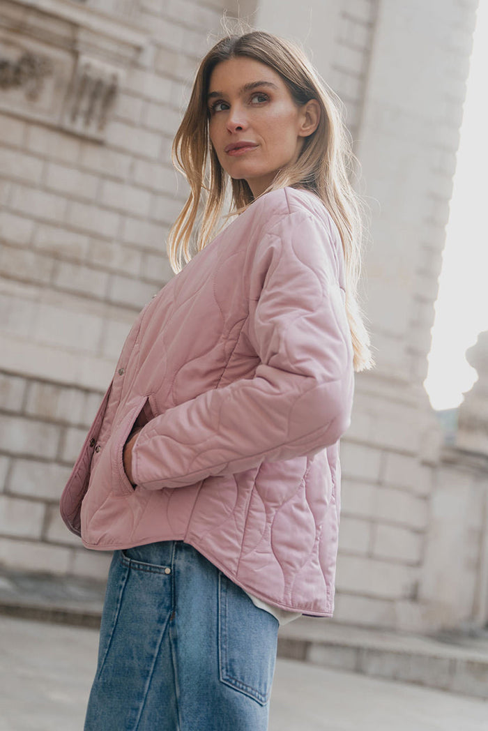 Woman wearing a pink quilted jacket standing in front of a stone building.