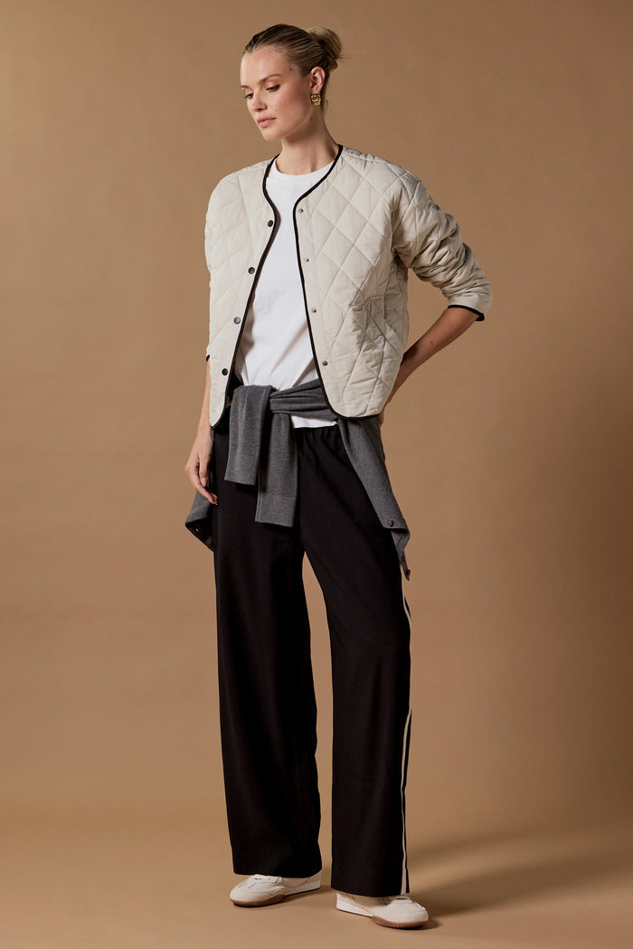 Woman wearing a beige quilted jacket, white shirt, gray cardigan, and black pants on a brown background