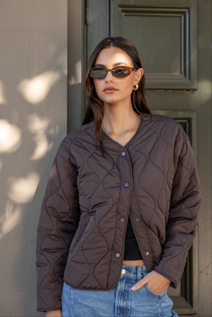 Woman wearing a brown quilted jacket and sunglasses standing against a neutral wall.
