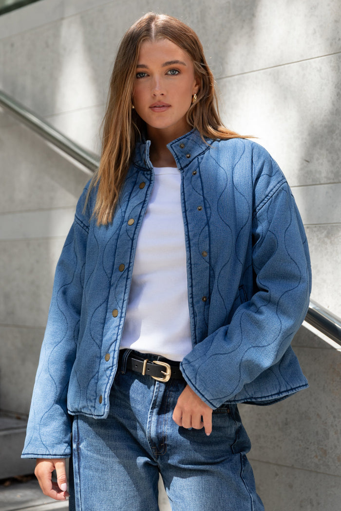Woman wearing a blue denim jacket and jeans standing against a gray wall.