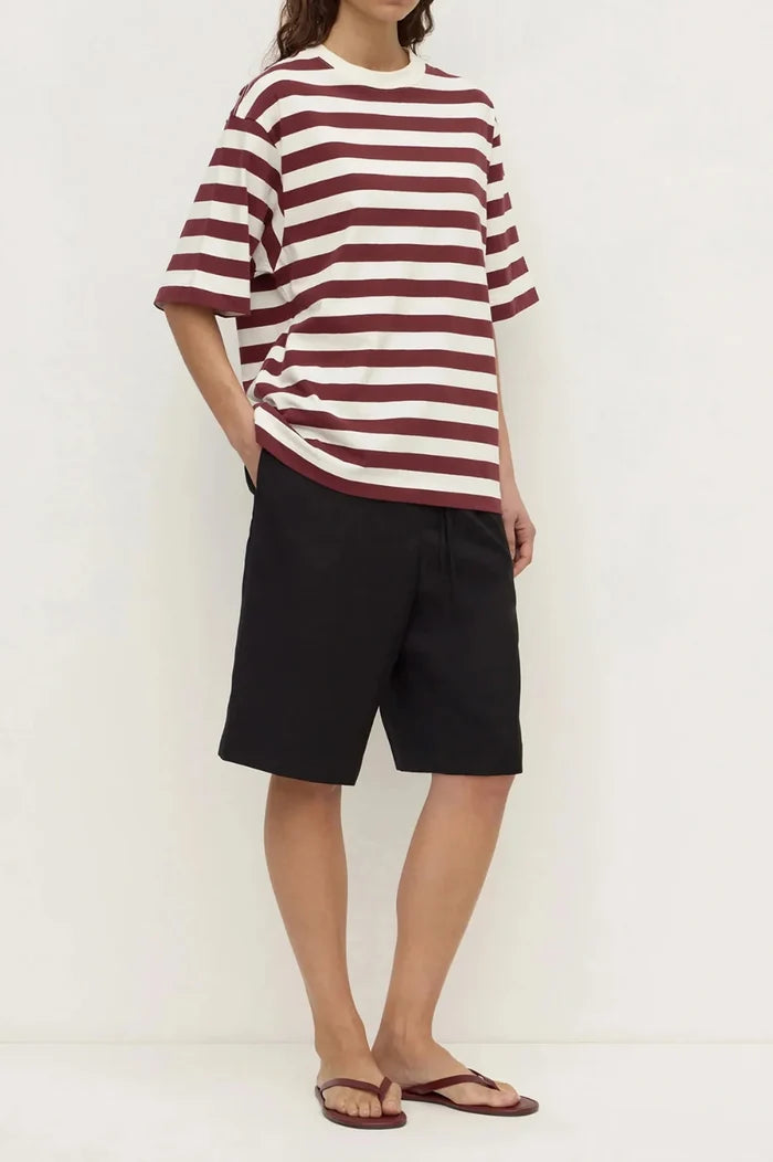 A model wearing a loose-fitting short-sleeve t-shirt with horizontal burgundy and off-white stripes, paired with black shorts and brown flip-flop sandals, standing against a plain white wall.