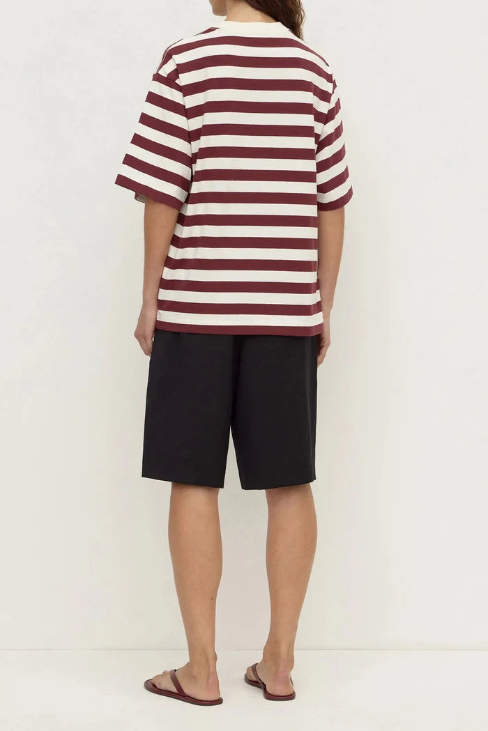 A person shown from the back wearing a short-sleeve striped t-shirt with burgundy and off-white horizontal stripes, paired with black shorts and brown sandals, against a plain off-white studio background.
