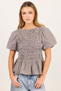 A woman modeling a short-sleeve smocked blouse with a subtle gray and white gingham pattern, paired with light-wash blue jeans, against a plain off-white studio background.