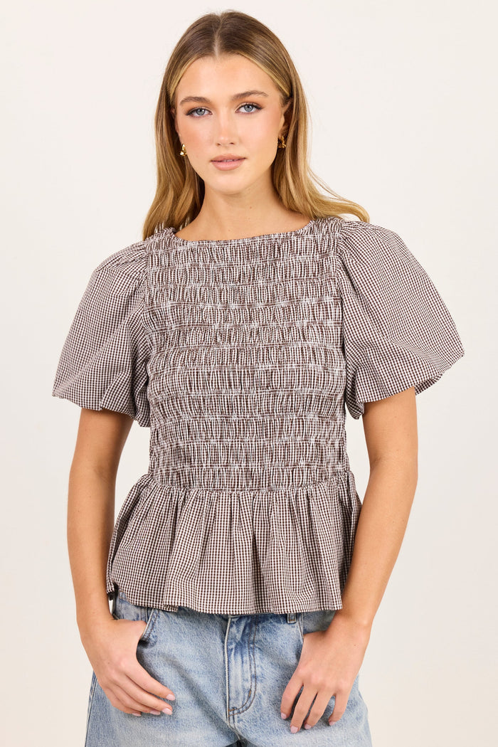 A woman modeling a short-sleeve smocked blouse with a subtle gray and white gingham pattern, paired with light-wash blue jeans, against a plain off-white studio background.
