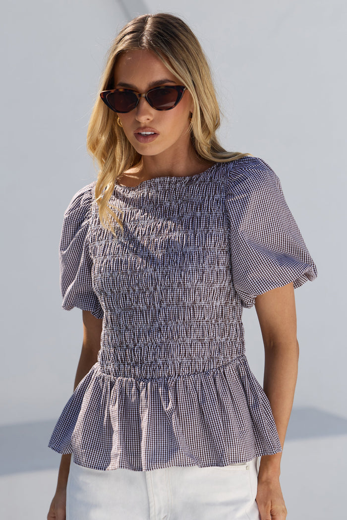 A woman wearing a chocolate and white gingham smocked blouse with puff sleeves and a peplum hem, paired with white pants, photographed against a clean white background.