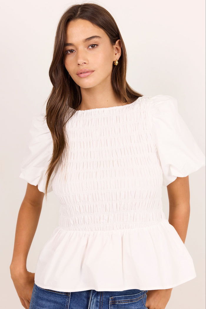A woman wearing a white smocked short-sleeve blouse with puff sleeves and a peplum hem, paired with blue jeans, against a plain off-white background.