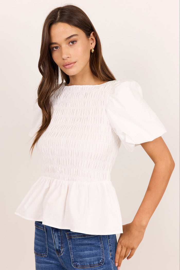 A woman wearing a white smocked blouse with puff sleeves and a peplum hem, paired with blue jeans, against a plain off-white background.