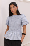 A woman modeling a blue and white vertically striped peplum blouse with puff sleeves, paired with black trousers, in an elegant indoor setting with white walls and molding.